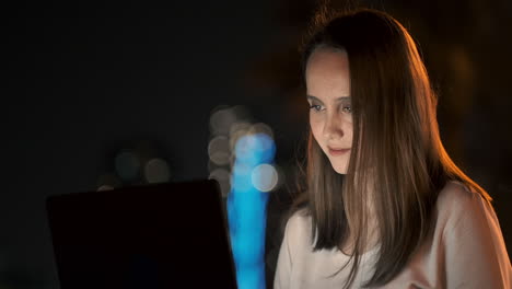 A-woman-software-developer-sits-in-a-Park-on-a-summer-night-in-the-city-and-writes-code-looking-at-a-laptop-screen.-Print-the-article-on-a-laptop.-Blogger-replies-to-posts